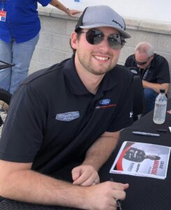 NASCAR driver Chase Briscoe smiles at the camera. He is wearing a black shirt and grey baseball cap and sunglasses.